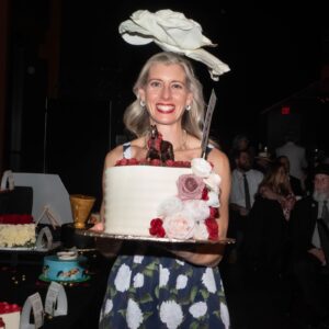 A blond woman wearing a blue dress with white flowers and a flower hat holding a cake
