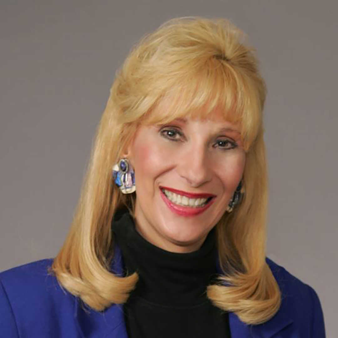 A white woman with light hair and a pink shirt stands in front of exotic trees