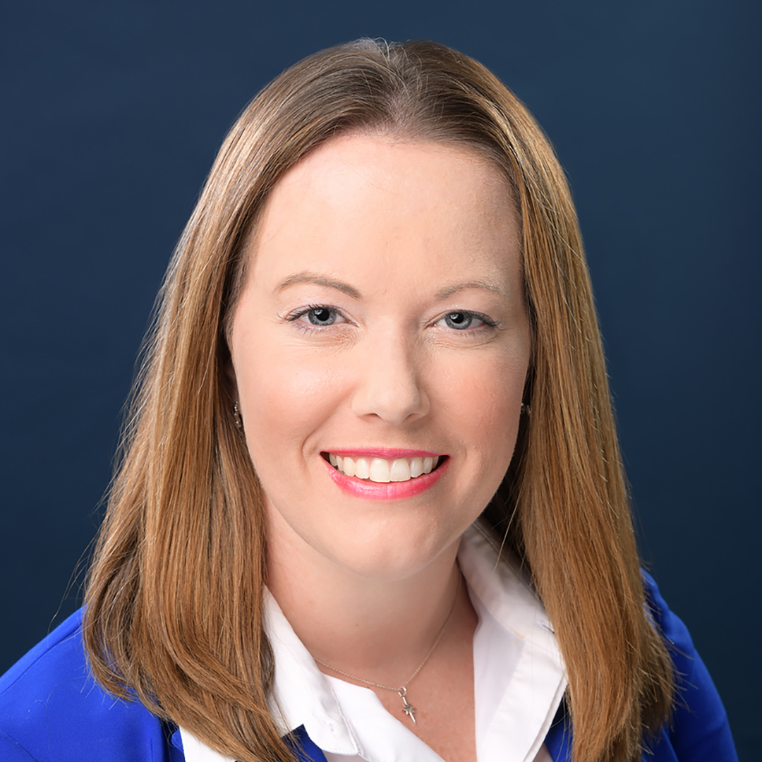 A white woman with light hair and a pink shirt stands in front of exotic trees