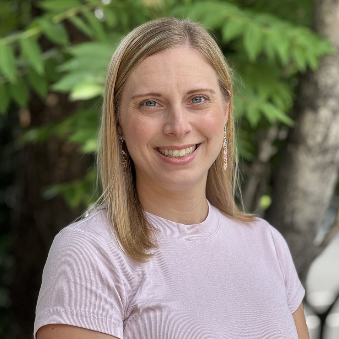 A white woman with light hair and a pink shirt stands in front of exotic trees