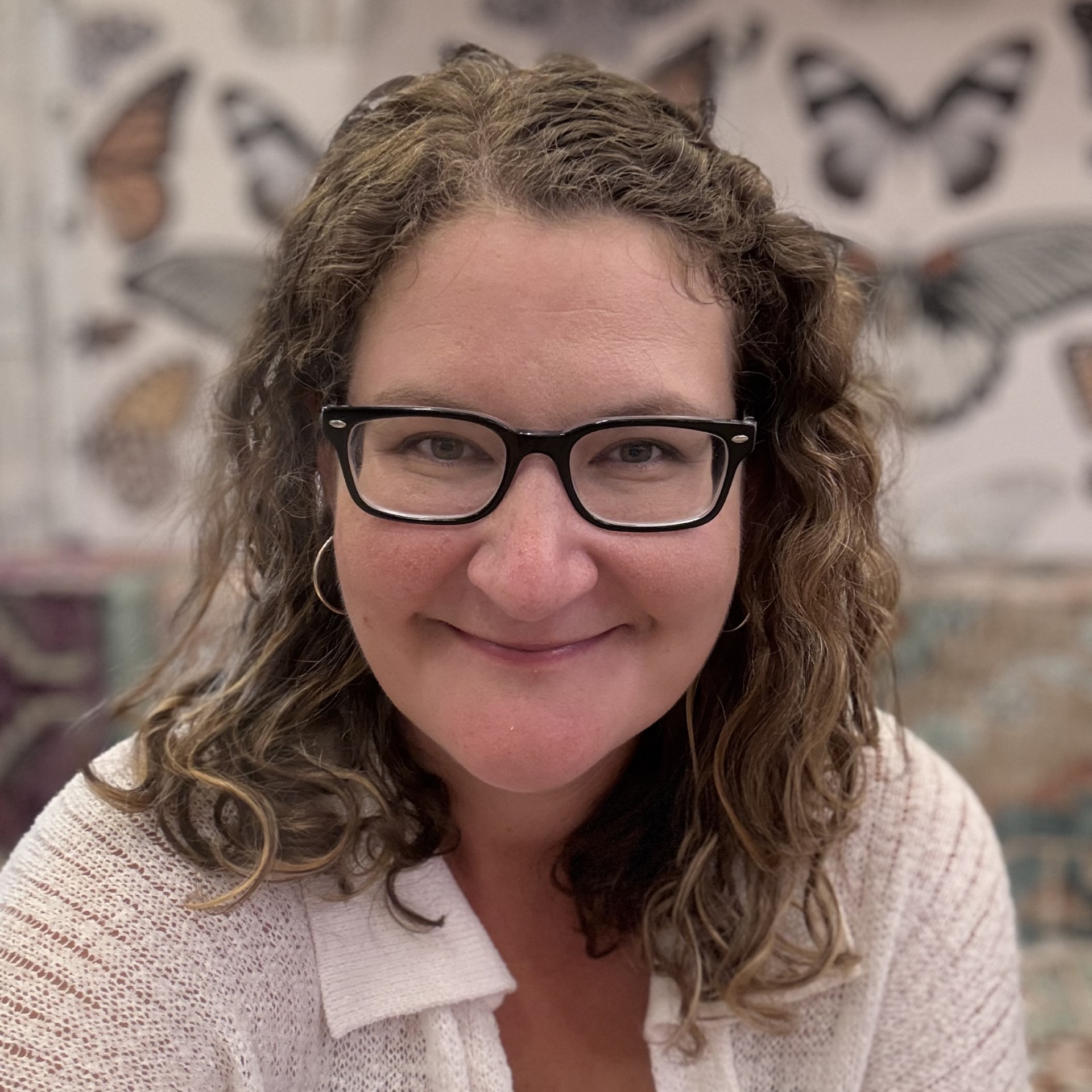 Theresa Kelly, a white woman with wavy brown hair and glasses in front of a wall with butterfly wallpaper