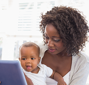 A black woman in a white shirt holds a black baby while reading a tablet