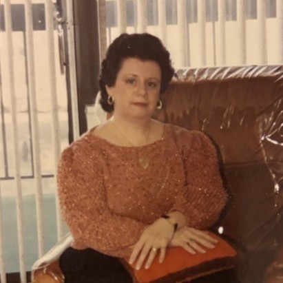 JoAnn Barbara Faitt-Jacobs, a middle aged woman with dark permed hair and a peach shirt sits on a leather chair in front of of a large window and blinds