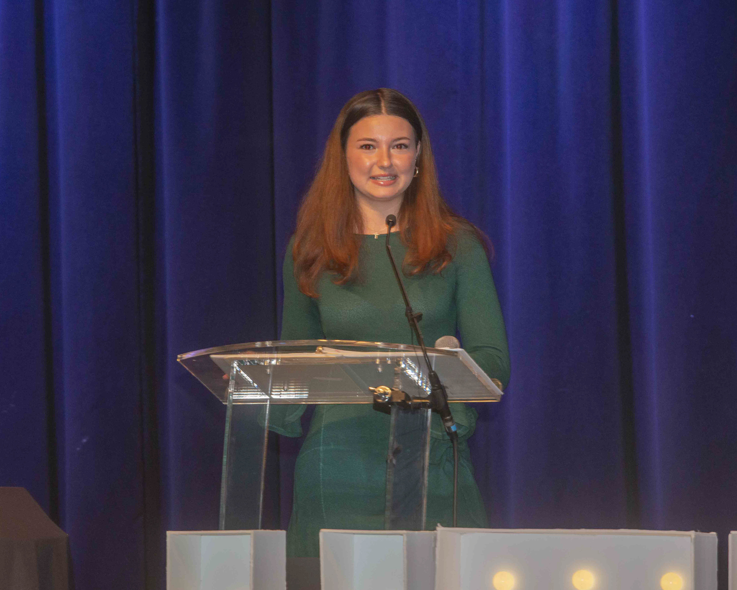 The 2026 honoree's granddaughter, a woman with red-ish hair, stands at a clear podium to accept the award.