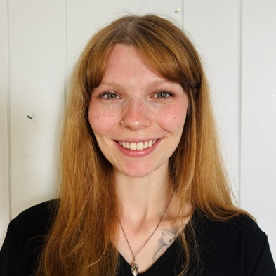 Audrey French, a white woman with long red hair smiles in front of a white paneled wall