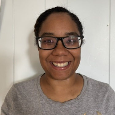 Joy Rodriguez, a dark skinned woman with short dark hair and glasses smiles against a white paneled background