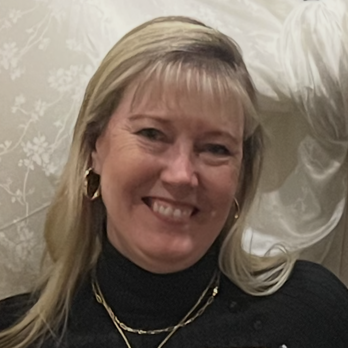 Margie Skibinski, a middle aged white woman with medium blond hair smiles in front of a grey wallpapered background